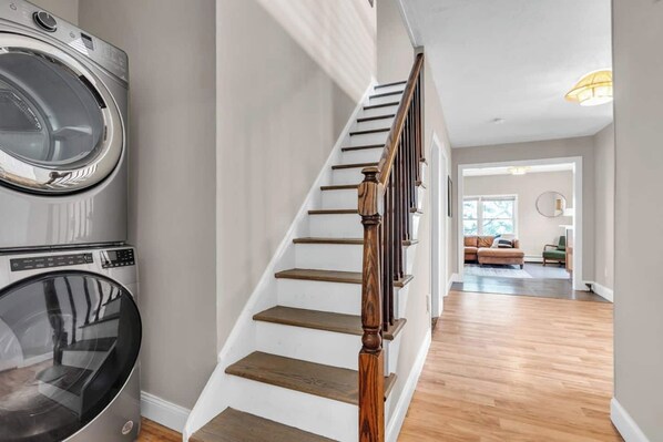 A laundry area with stacked washers and dryers, a wooden staircase, and a living room with a sofa and a lamp.