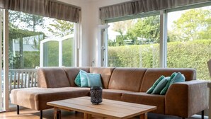 Una sala de estar moderna con un sofá marrón, una mesa de centro de madera y grandes ventanales que dan a vistas de vegetación.