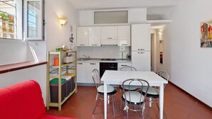 A compact kitchen with white cabinets, a dining table with four chairs, a red sofa, and a window with a view of buildings.