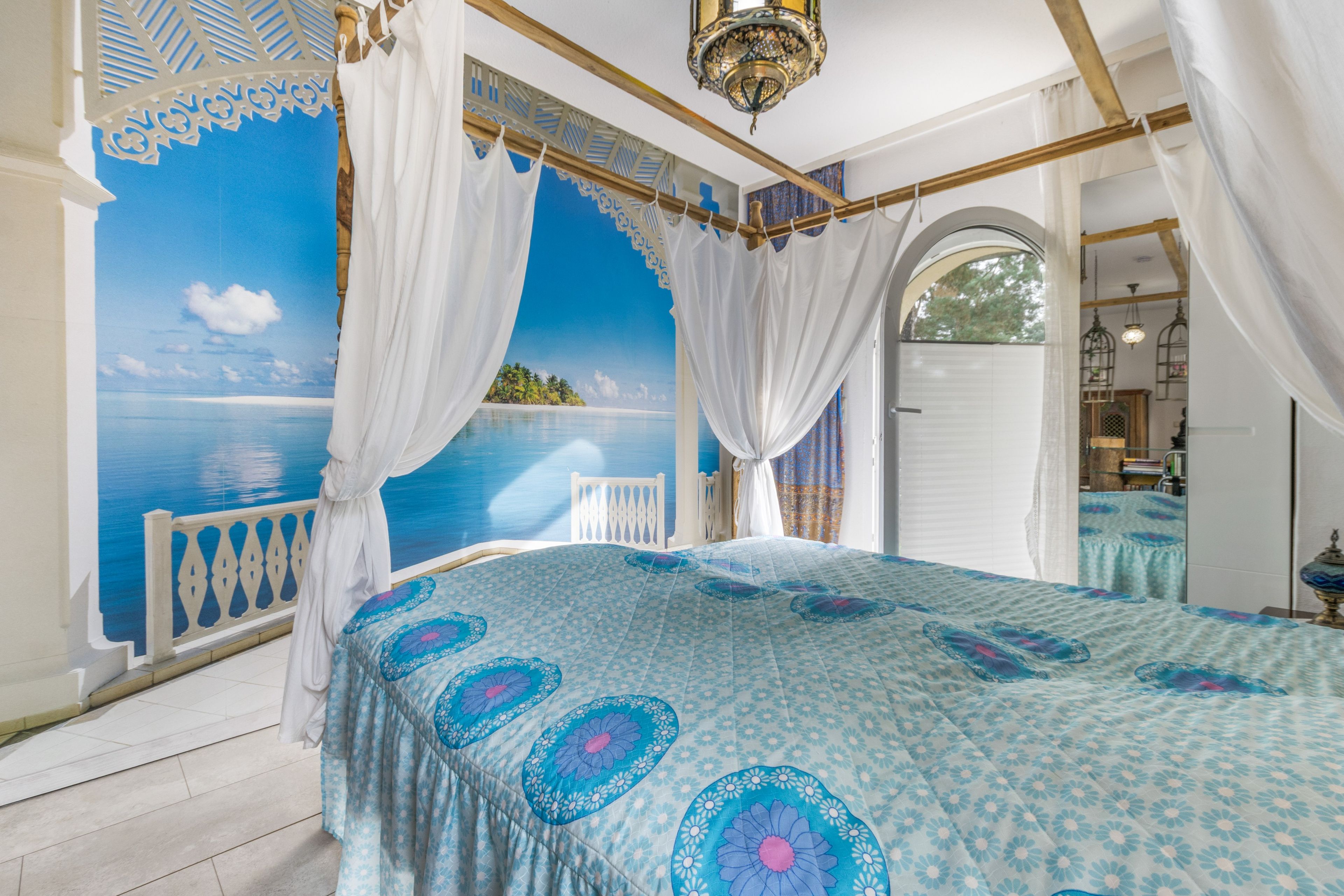 A bedroom with a large bed, a view of the sea through a window, and a balcony with a white railing.