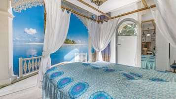A bedroom with a large bed, a view of the sea through a window, and a balcony with a white railing.