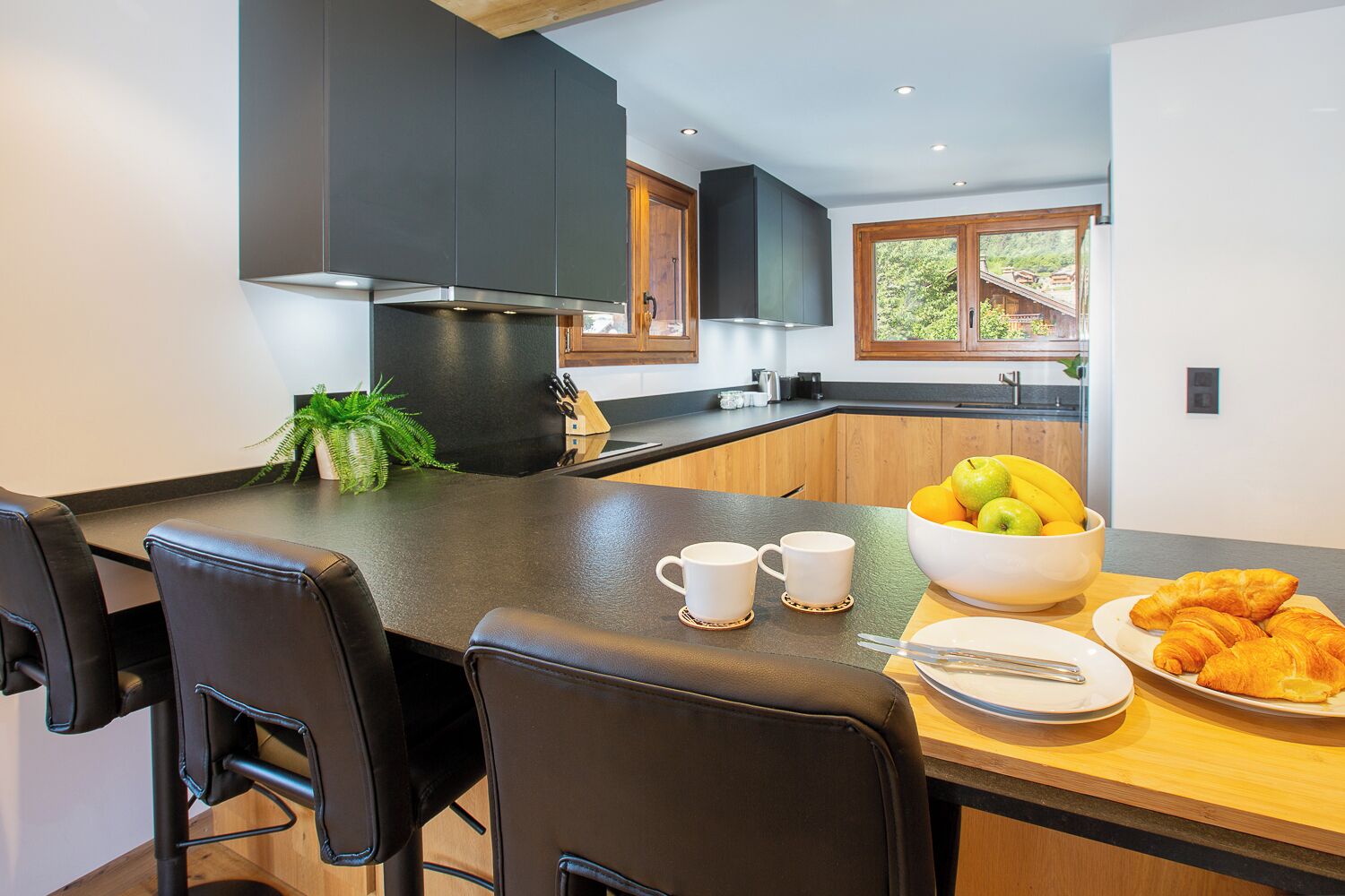Een moderne keuken met een centraal eiland, zwarte stoelen, een schaal fruit en croissants op het aanrecht.