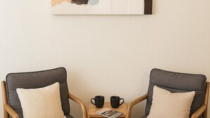 Zwei graue Sessel mit Holzgestell und beigen Kissen, ein kleiner Holztisch mit zwei schwarzen Tassen und ein großes abstraktes Gemälde an der Wand.