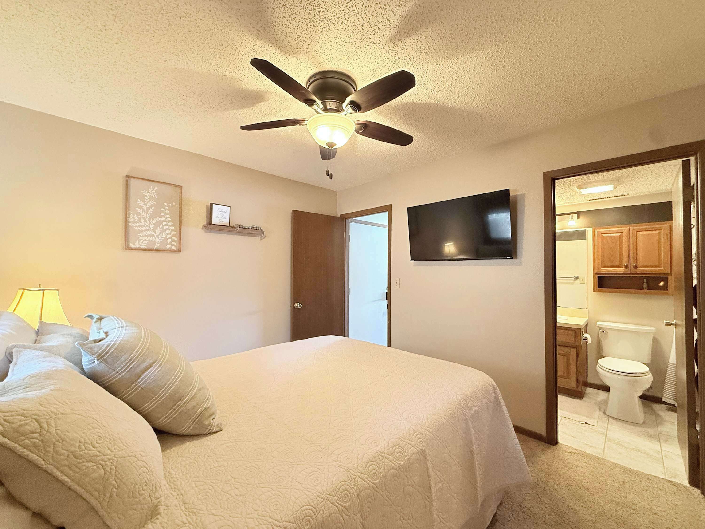 A bedroom with a bed, a ceiling fan, a TV, and a door leading to a bathroom.