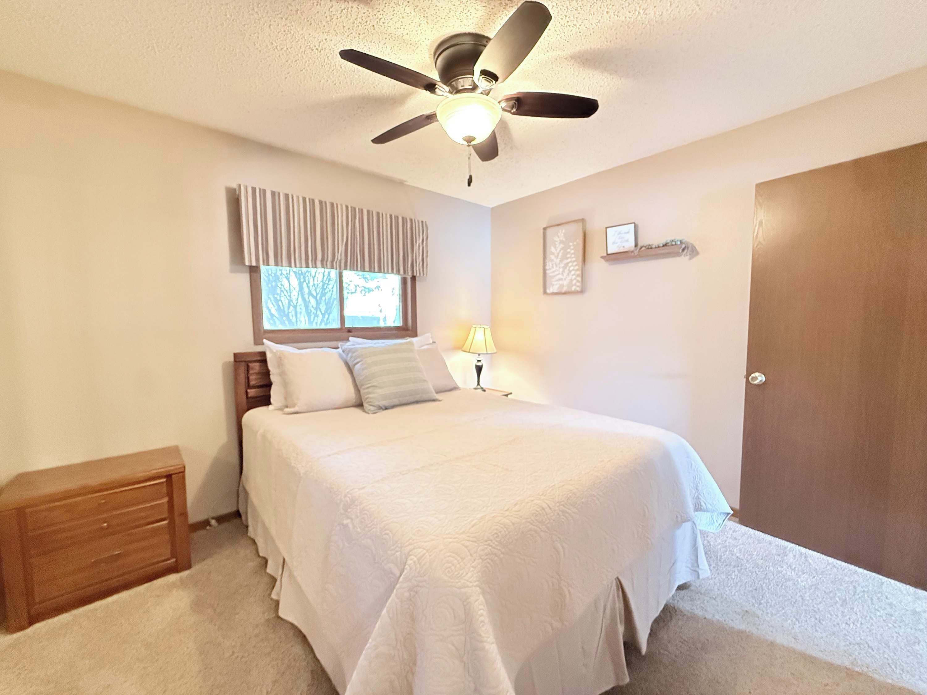 A bedroom with a bed, a ceiling fan, a window with curtains, a bedside lamp, a wooden nightstand, and a door.
