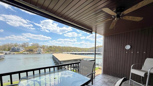 A balcony with a glass table and chairs, a ceiling fan, and a view of a lake with buildings in the background. - Loto fun at 21 Lakeview Condo (Osage Beach)