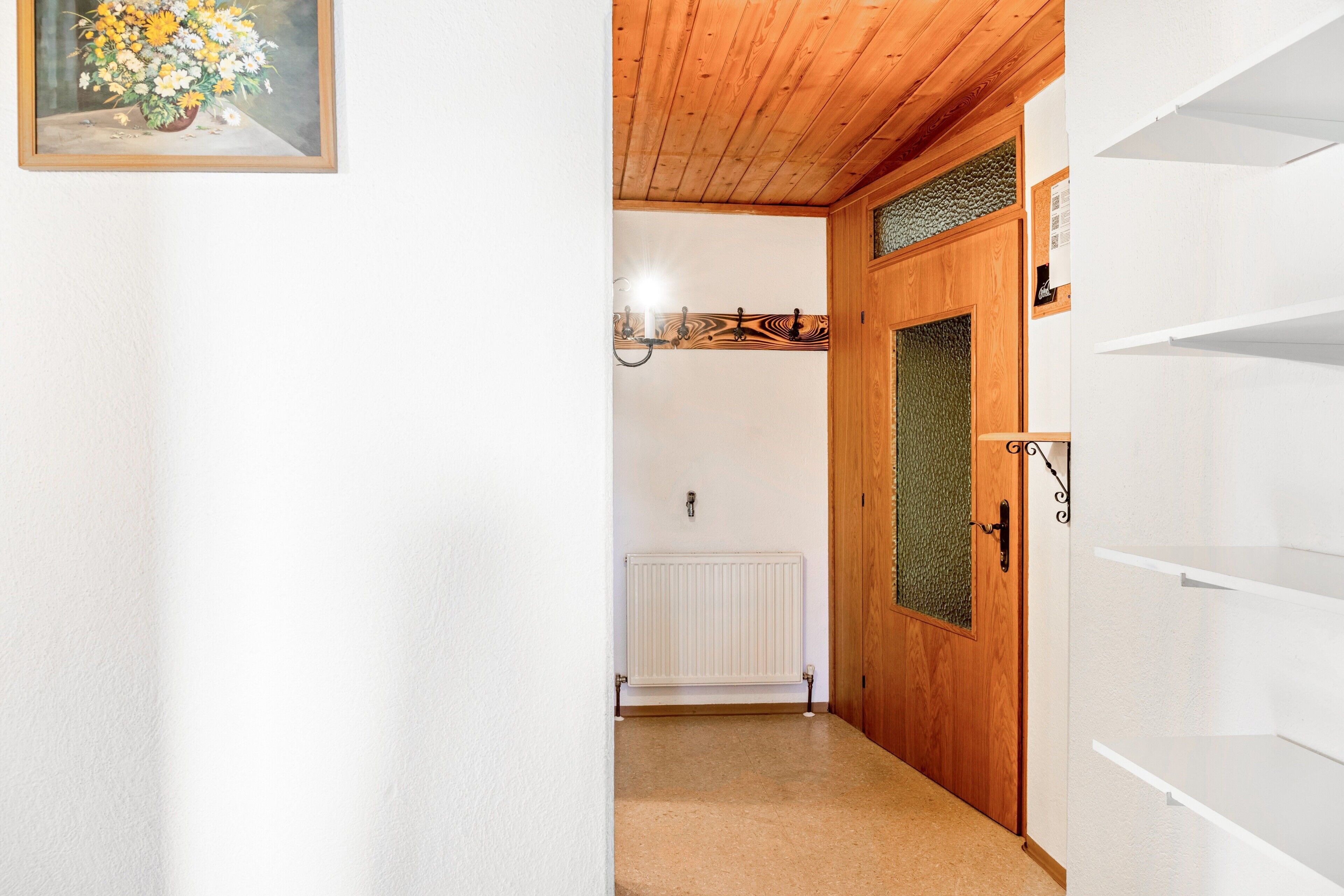 Een gang met een houten plafond, een radiator, een deur met ruit en een lijstwerk met een afbeelding aan de muur.