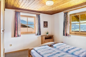 Une chambre avec un plafond en bois, deux fenêtres avec des rideaux, un lit recouvert d’une couverture à carreaux bleus et blancs, une table de chevet en bois et une petite plante en pot.