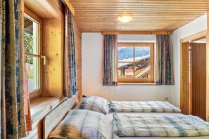 Une pièce avec un plafond en bois, une fenêtre avec des rideaux, un lit recouvert d’une couverture à carreaux, et une vue sur une montagne et un bâtiment à l’extérieur.