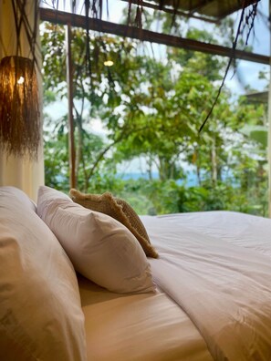 Ein Bett mit weißer Bettwäsche und einem Leinenkissen, eine hängende Leuchte und Blick ins Grüne durch ein Fenster.