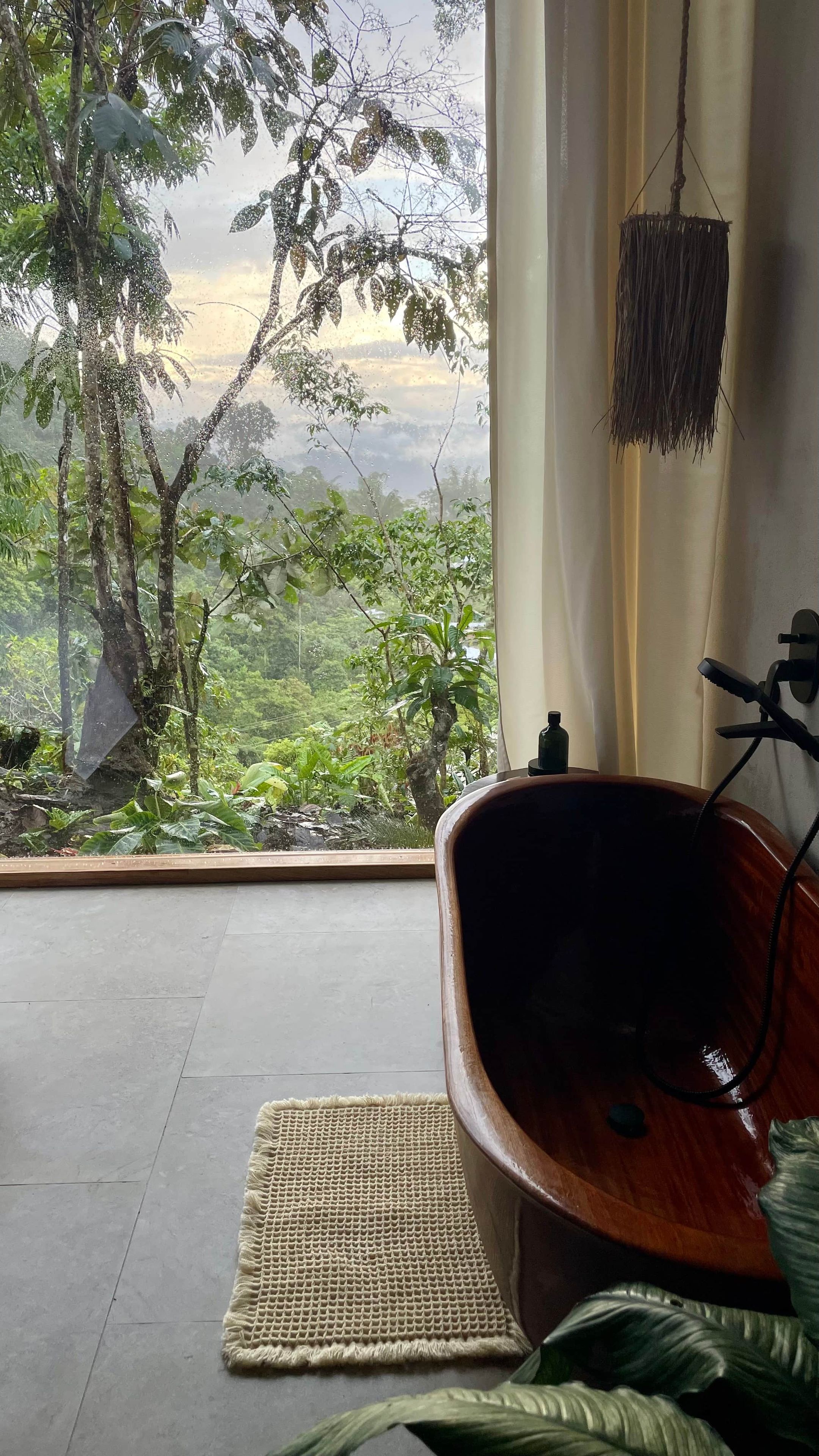 A room with a view of a forest, a wooden bathtub, and a woven mat on the floor.