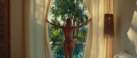 A room with a bed, a window with sheer curtains, and a view of greenery outside.