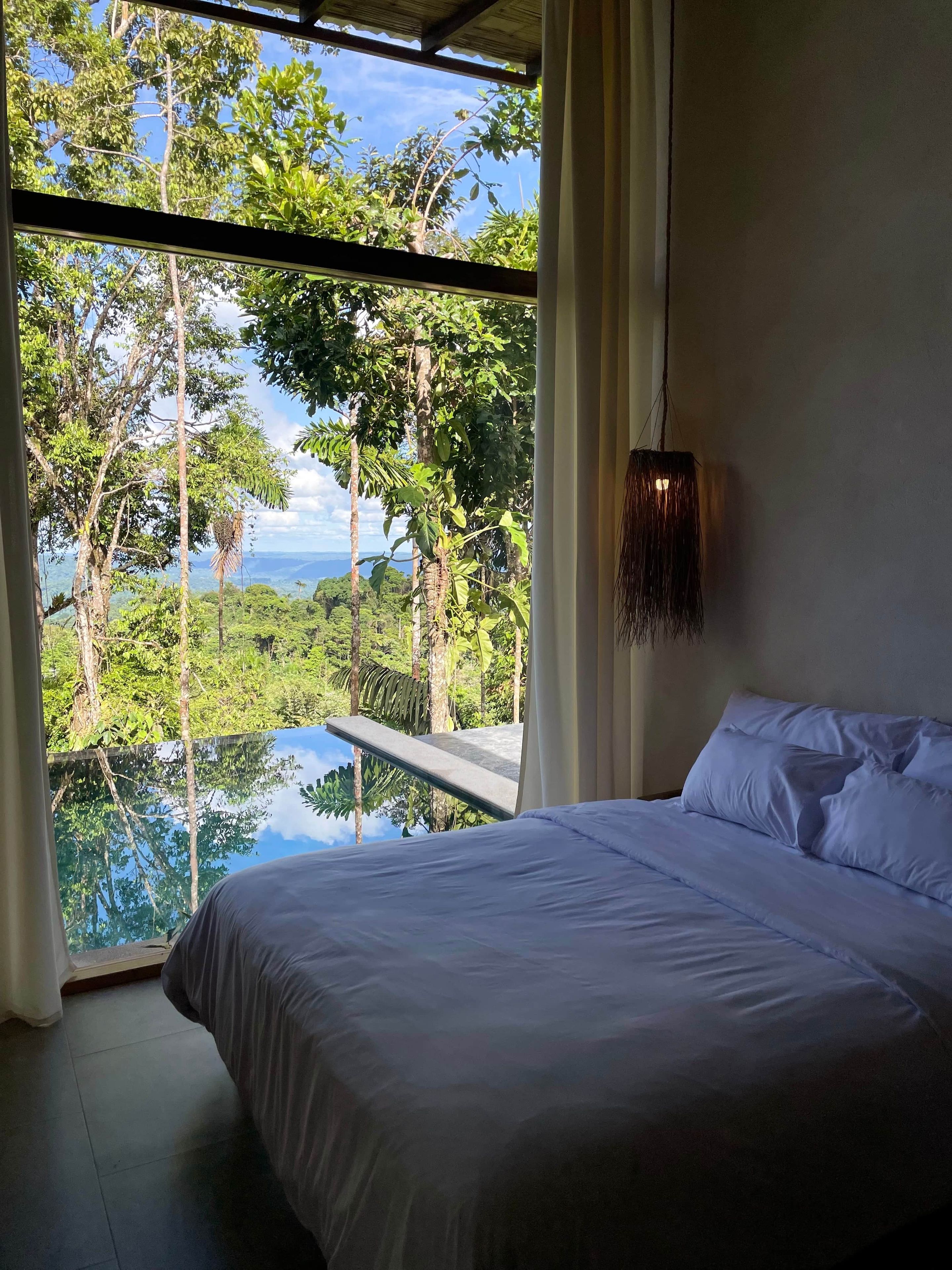 A bedroom with a large bed, a view of a lush landscape, and a hanging light fixture.
