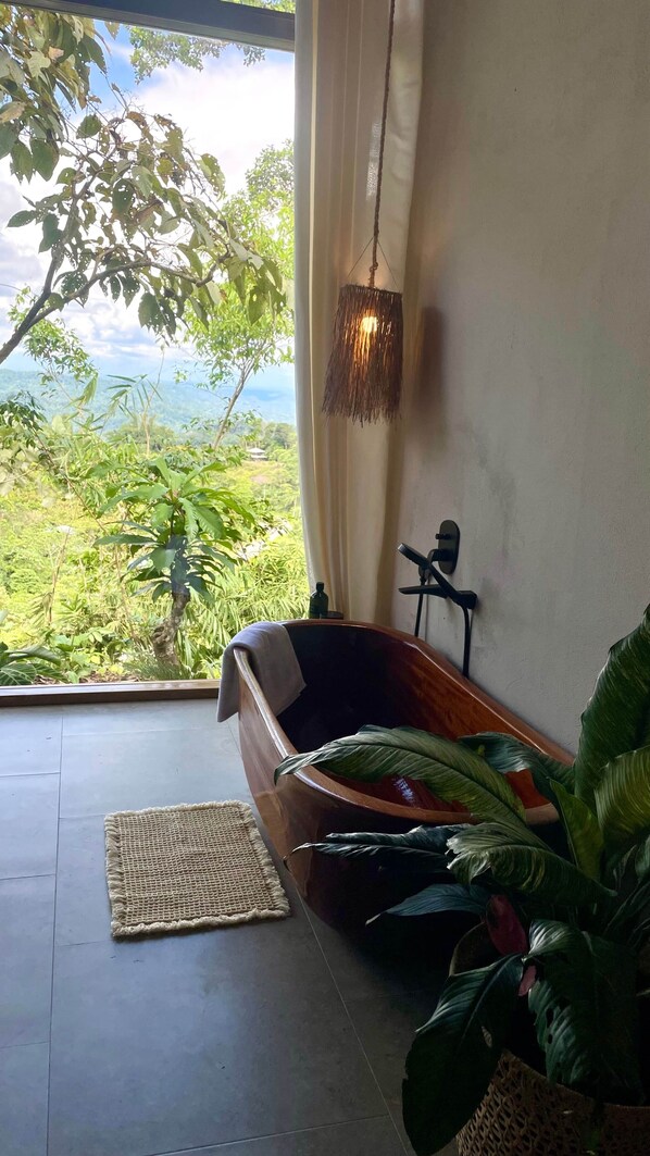 A bathroom with a wooden tub, a hanging light fixture, and a view of greenery outside.