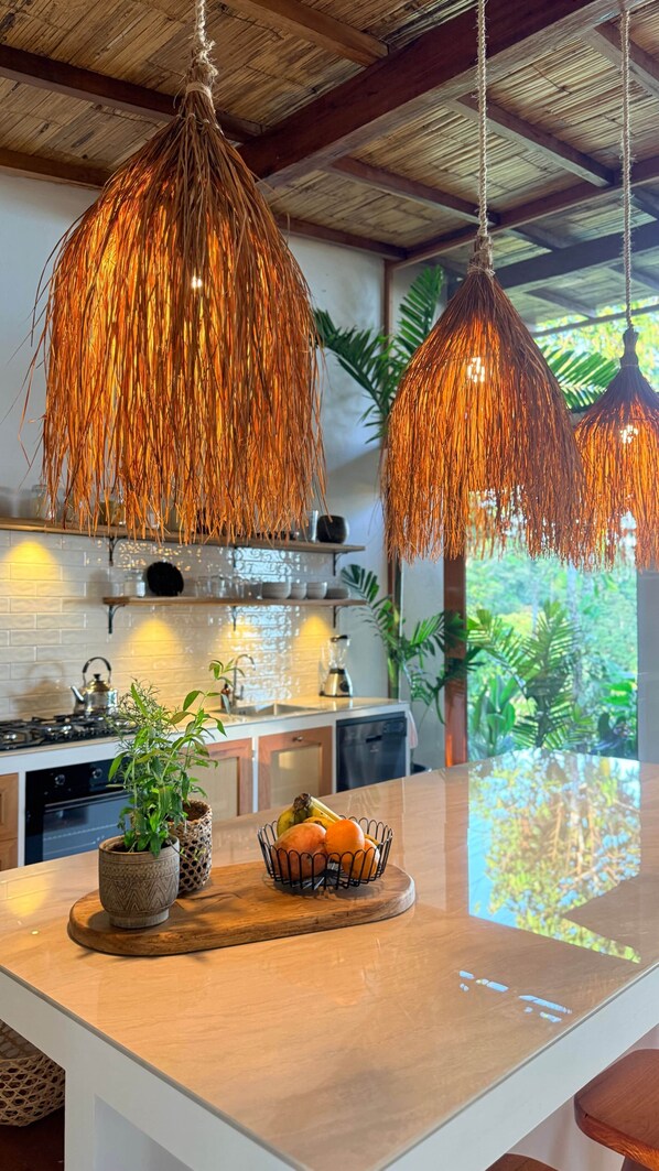 Une cuisine dotée de luminaires suspendus, d’un îlot central agrémenté d’un panier de fruits et offrant une vue sur la verdure à l’extérieur.