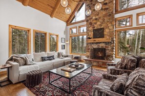 A cozy living room with a stone fireplace, large windows, and a high wooden ceiling. - Exclusive Pet-Friendly Mountain Escape | Arcade, Hot Tub, Views & Steps to Ski (Mountain Village)