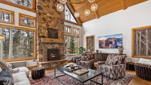 Un salon chaleureux avec une cheminée en pierre, de grandes fenêtres et un haut plafond en bois.