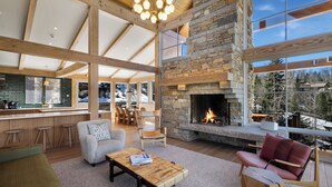 A spacious living area with a stone fireplace, wooden beams, and large windows offering a view of the outdoors. - Ski In/Out, Hot tub, Sauna, Fireplaces, Fire Pit, Game Room (Mountain Village)