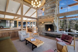 A spacious living area with a stone fireplace, wooden beams, and large windows offering a view of the outdoors. - Ski In/Out, Hot tub, Sauna, Fireplaces, Fire Pit, Game Room (Mountain Village)