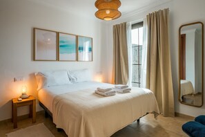 Ein Hotelzimmer mit Bett, Nachttischlampen, einem Fenster mit Vorhängen, einem Wandspiegel und gerahmten Kunstwerken.