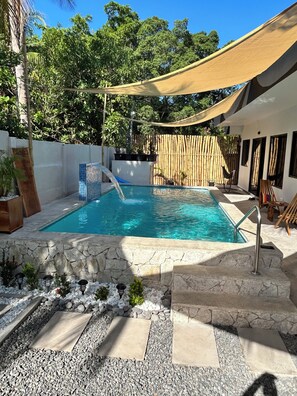 En swimmingpool med stenkant, bambushegn og et parasolsejl.