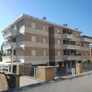A multi-story residential building with balconies, a visible ground-level entrance, and a clear sky. - House Olgiata (Roma)