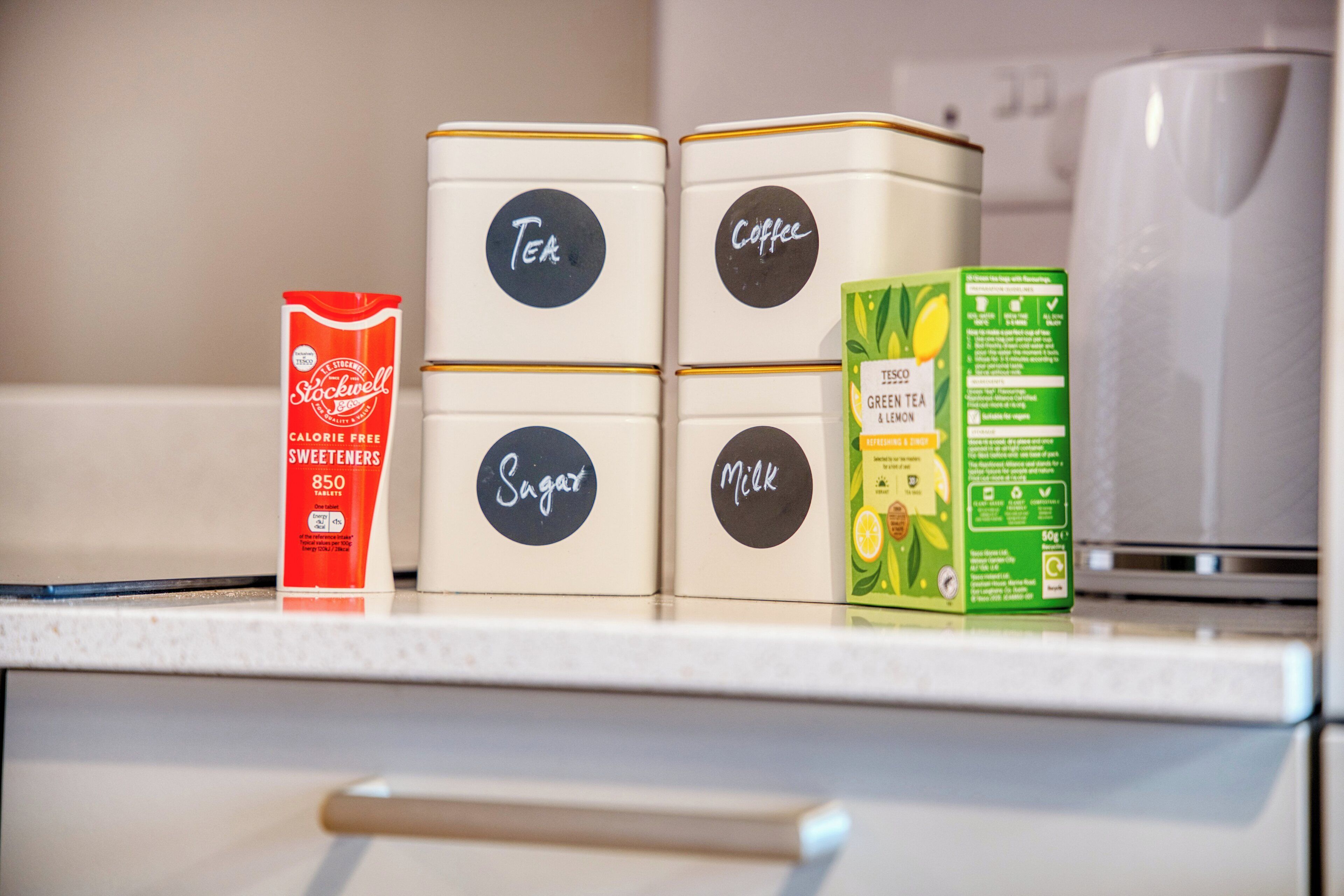 Vier gelabelde containers op een aanrecht: Thee, Koffie, Suiker en Melk.