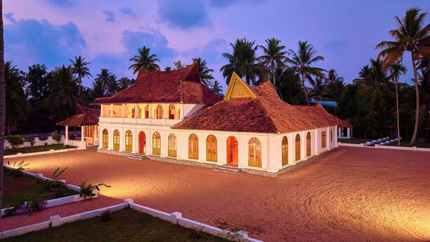 A white building with red-tiled roofs, arched windows, and a paved courtyard surrounded by palm trees.