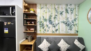 A compact living space with a kitchenette, a sofa with cushions, a wall-mounted shelf with decorative items, and a bamboo-themed wallpaper. - Sunset View 3-Bedroom SMDC (Pasay)