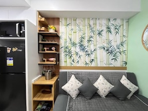A compact living space with a kitchenette, a sofa with cushions, a wall-mounted shelf with decorative items, and a bamboo-themed wallpaper. - Sunset View 3-Bedroom SMDC (Pasay)