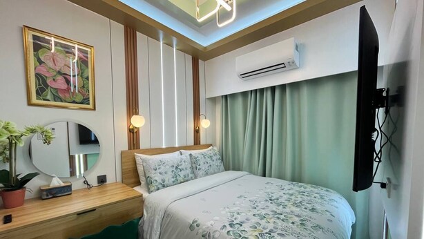 A hotel room with a bed, desk, chair, bedside table, mirror, potted plant, and an air conditioning unit. - Sunset View 3-Bedroom SMDC (Pasay)