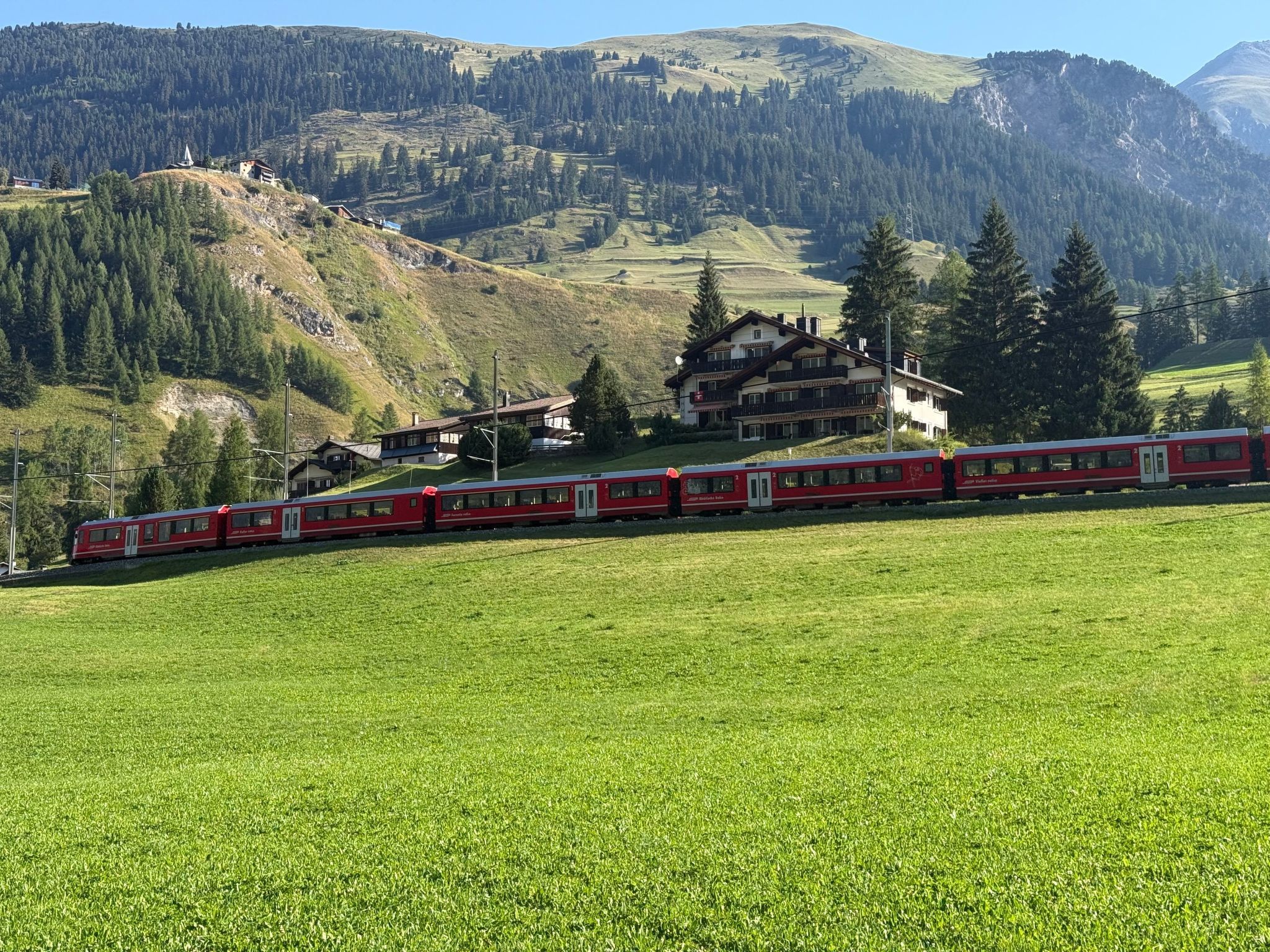 A red train on a track with a mountainous backdrop, a building with a sloped roof, and a green field in the foreground.