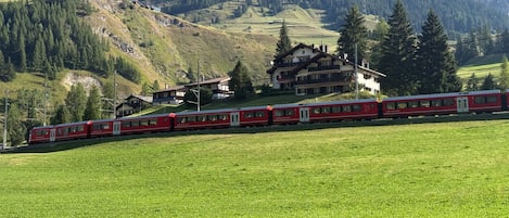 A red train on a track with a mountainous backdrop, a building with a sloped roof, and a green field in the foreground.