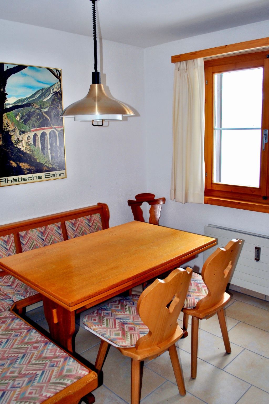 Un coin repas avec une table et des chaises en bois, un luminaire suspendu, une fenêtre avec des rideaux et un tableau accroché au mur.