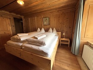 A wooden-framed bed with pillows and a blanket, a bedside table with a lamp, and a window with curtains. - Haus Buol Bergün, (Bergün/Bravuogn), 213 by Interhome (Bergün)