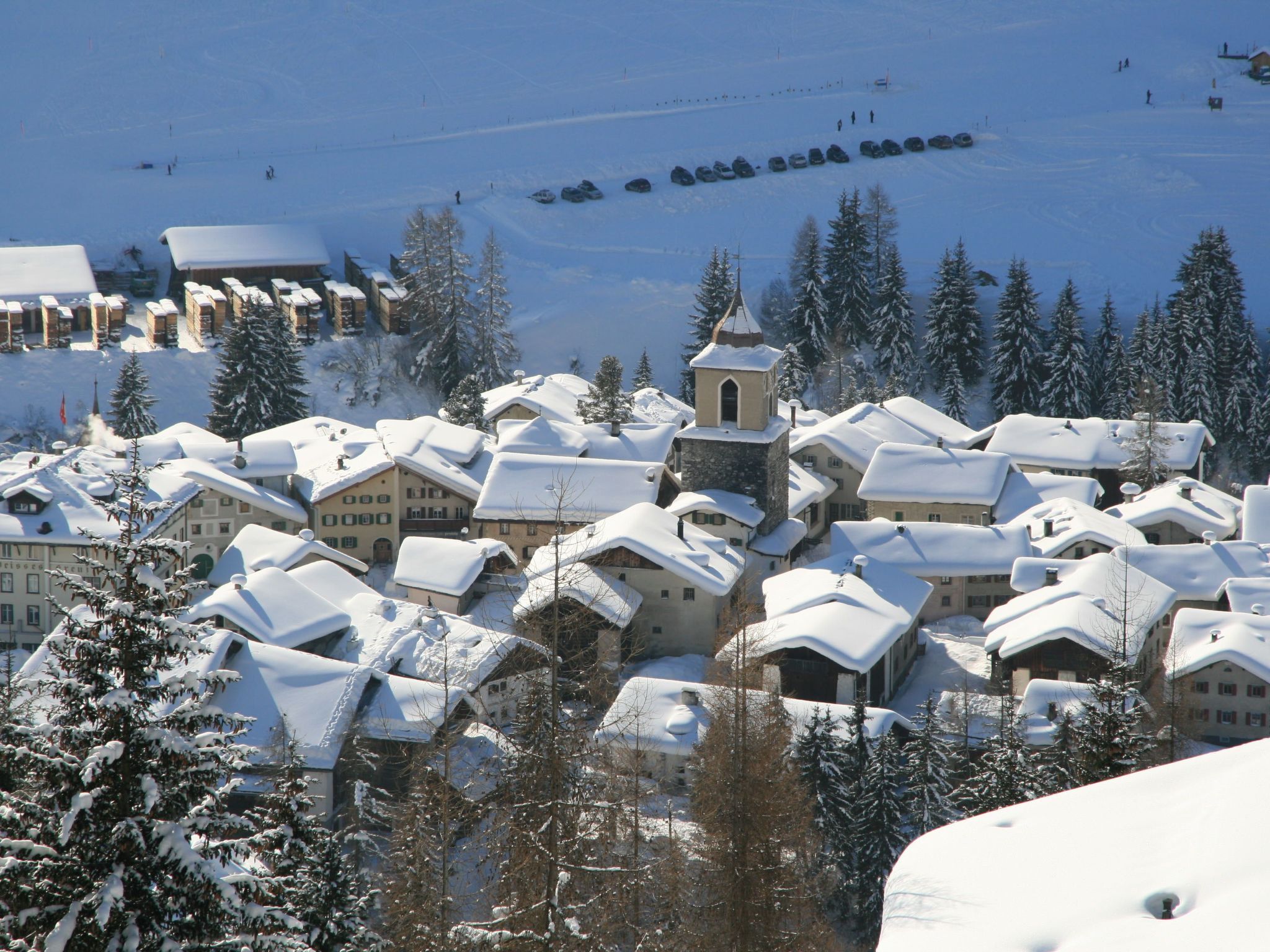 Ein schneebedecktes Dorf mit traditionellen Gebäuden, einer Kirche und einer Skipiste im Hintergrund.