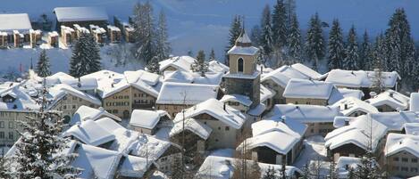 Ein schneebedecktes Dorf mit traditionellen Gebäuden, einer Kirche und einer Skipiste im Hintergrund.
