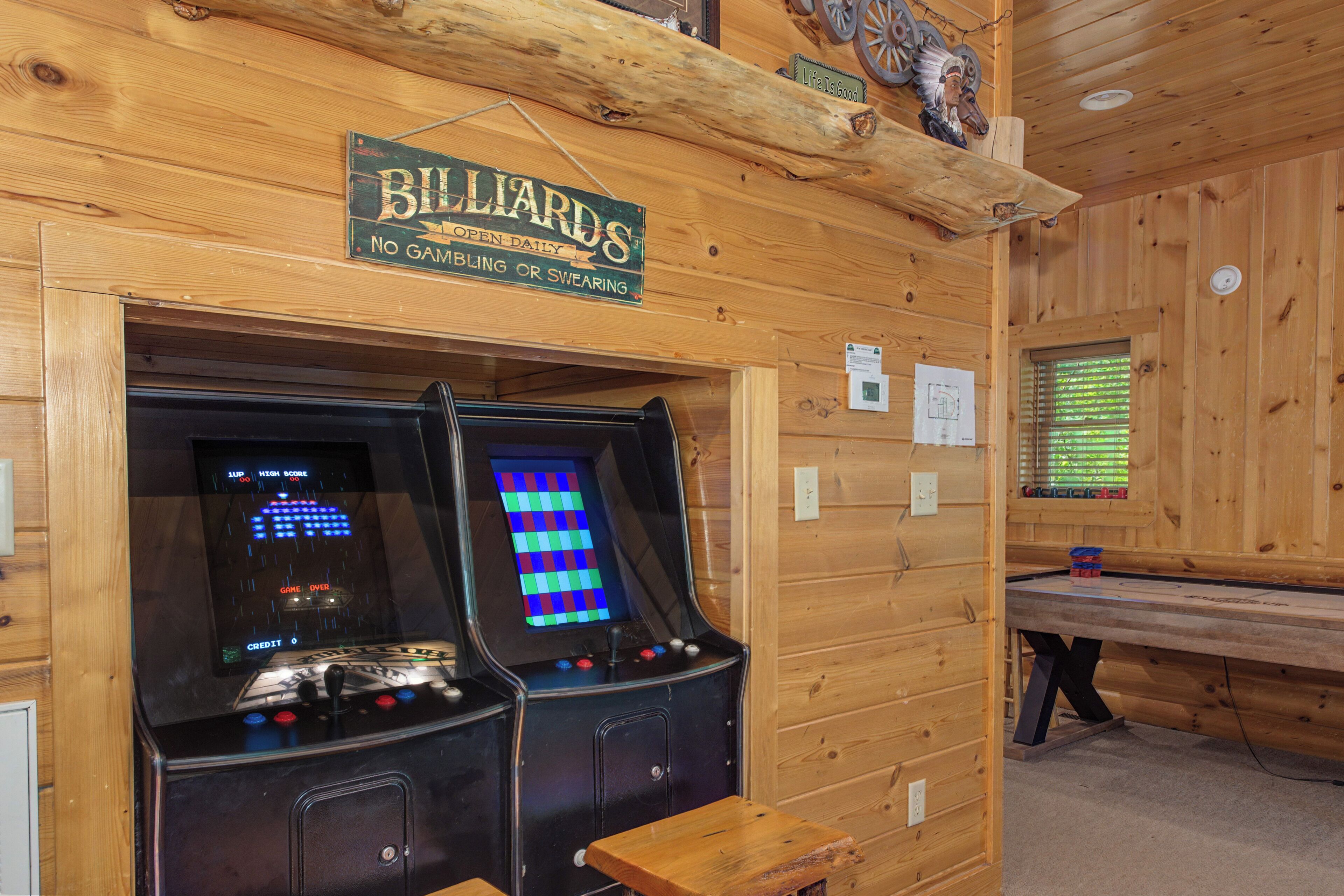 Un intérieur en bois avec deux bornes d’arcade, un panneau indiquant la présence d’un billard et une fenêtre avec des stores.