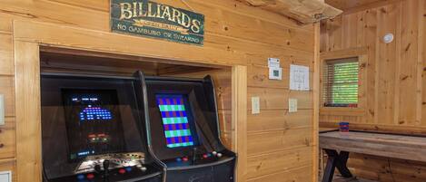 Un intérieur en bois avec deux bornes d’arcade, un panneau indiquant la présence d’un billard et une fenêtre avec des stores.