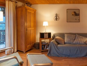 Ein gemütliches Wohnzimmer mit einem Holzschrank, einer Couch mit gemusterten Kissen, einem kleinen Tisch mit einer Lampe und einem Bücherregal.