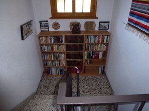 Un escalier menant à une bibliothèque remplie de livres, une fenêtre au-dessus, et un mur orné de tableaux encadrés.