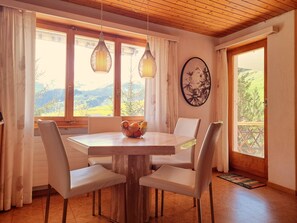Ein Essbereich mit einem runden Tisch, vier Stühlen, einer Obstschale und Bergblick durch große Fenster.
