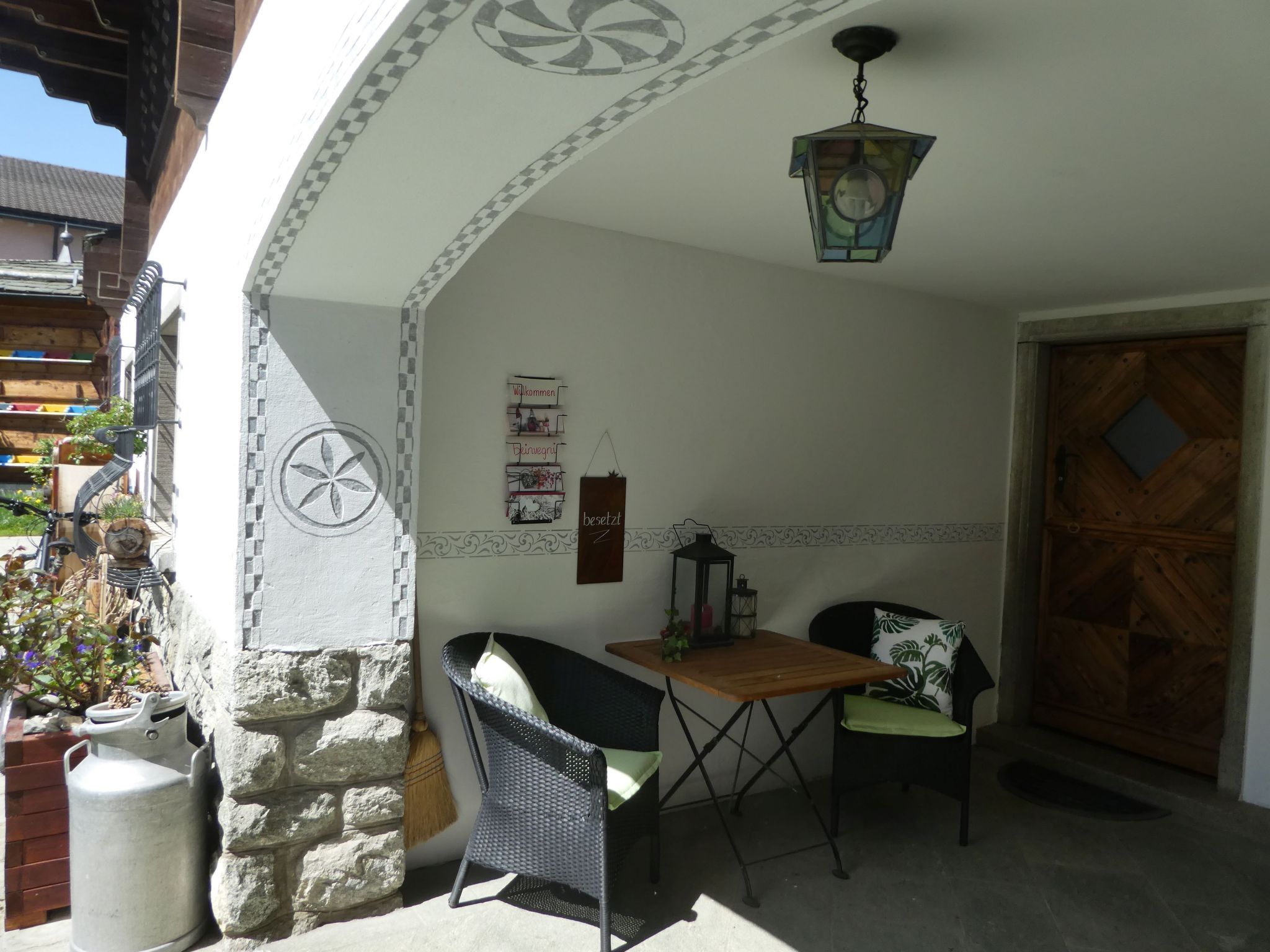 A small outdoor seating area with a table and two chairs, a lantern, and a decorative wall with a welcome sign.
