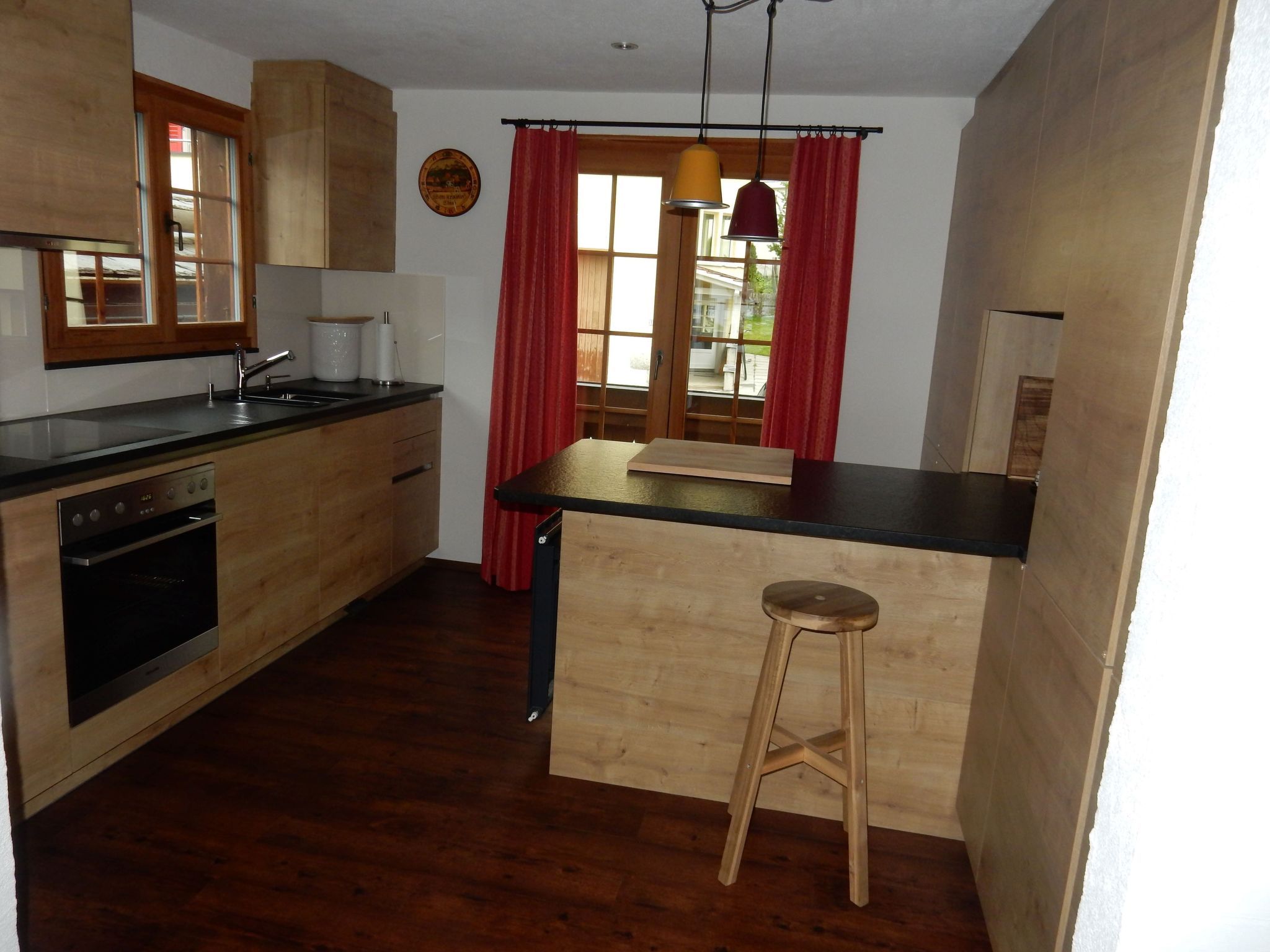 Een kleine keuken met houten kastjes, een inbouwoven, een spoelbak en een barstoel.