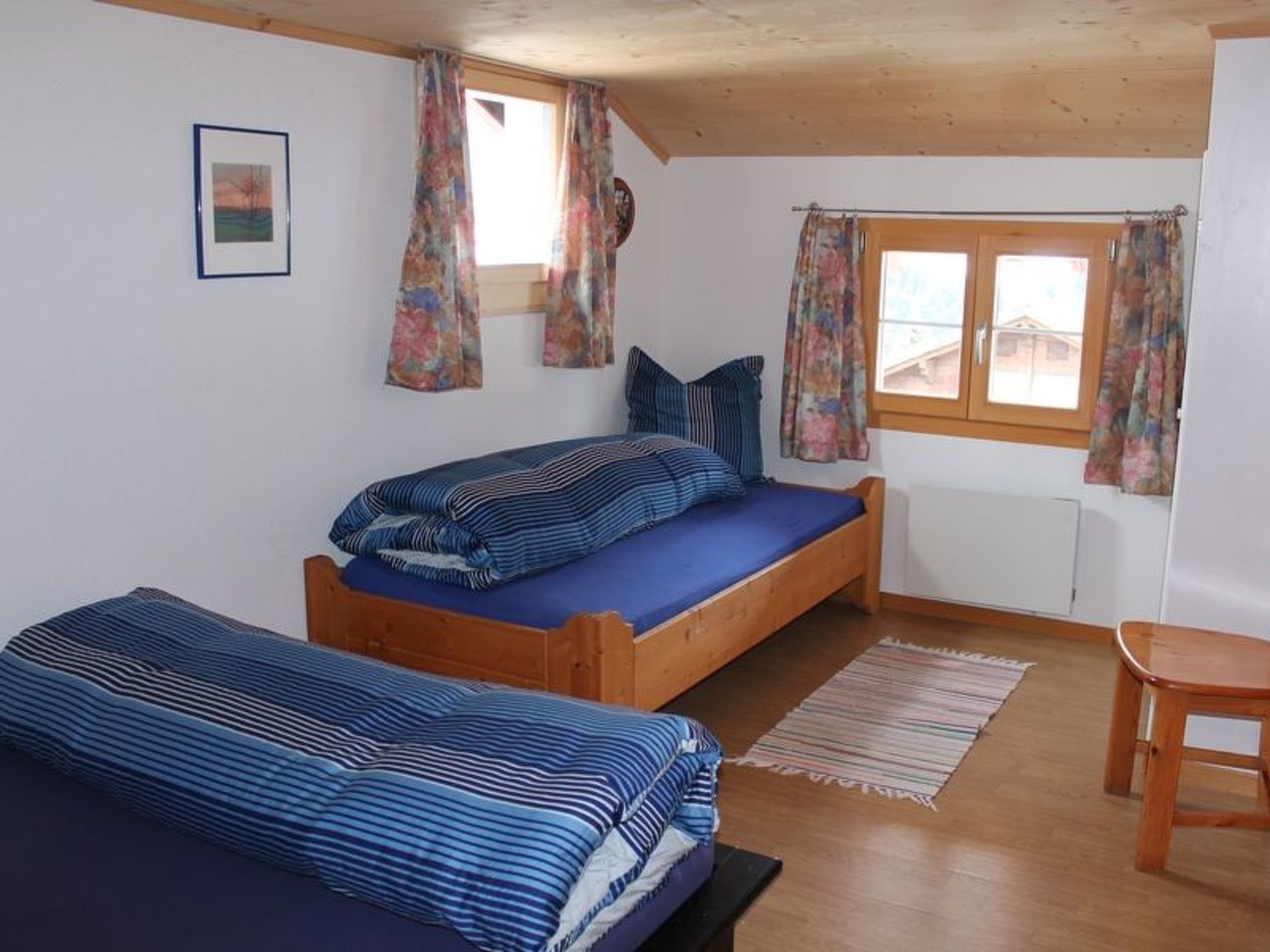 Une pièce avec deux lits en bois, une chaise en bois, une fenêtre avec des rideaux et un radiateur.