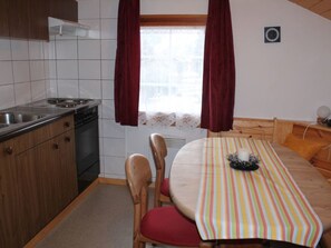Eine kompakte Küche mit Herd, Spüle und Schränken, ein Esstisch mit Stühlen sowie ein Fenster mit Vorhängen.