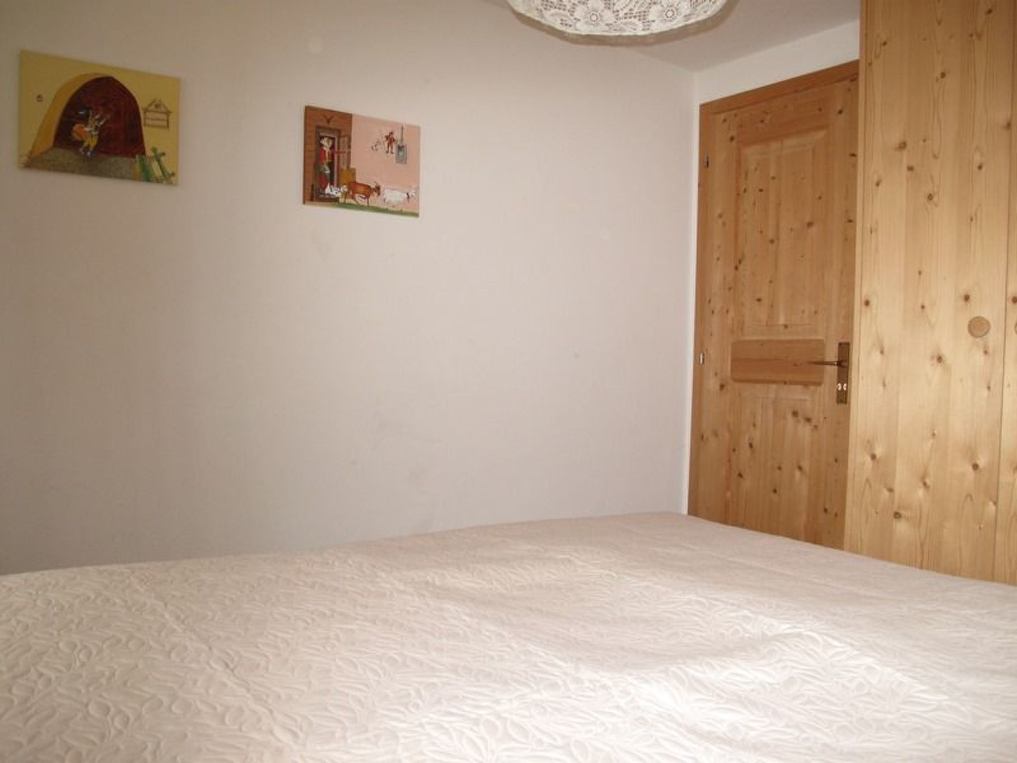 A bedroom with a wooden door, a bed with a white textured mattress, and two wall-mounted pictures.