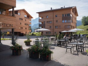 A modern building with multiple floors, outdoor seating area with tables and umbrellas, and a mountainous backdrop.