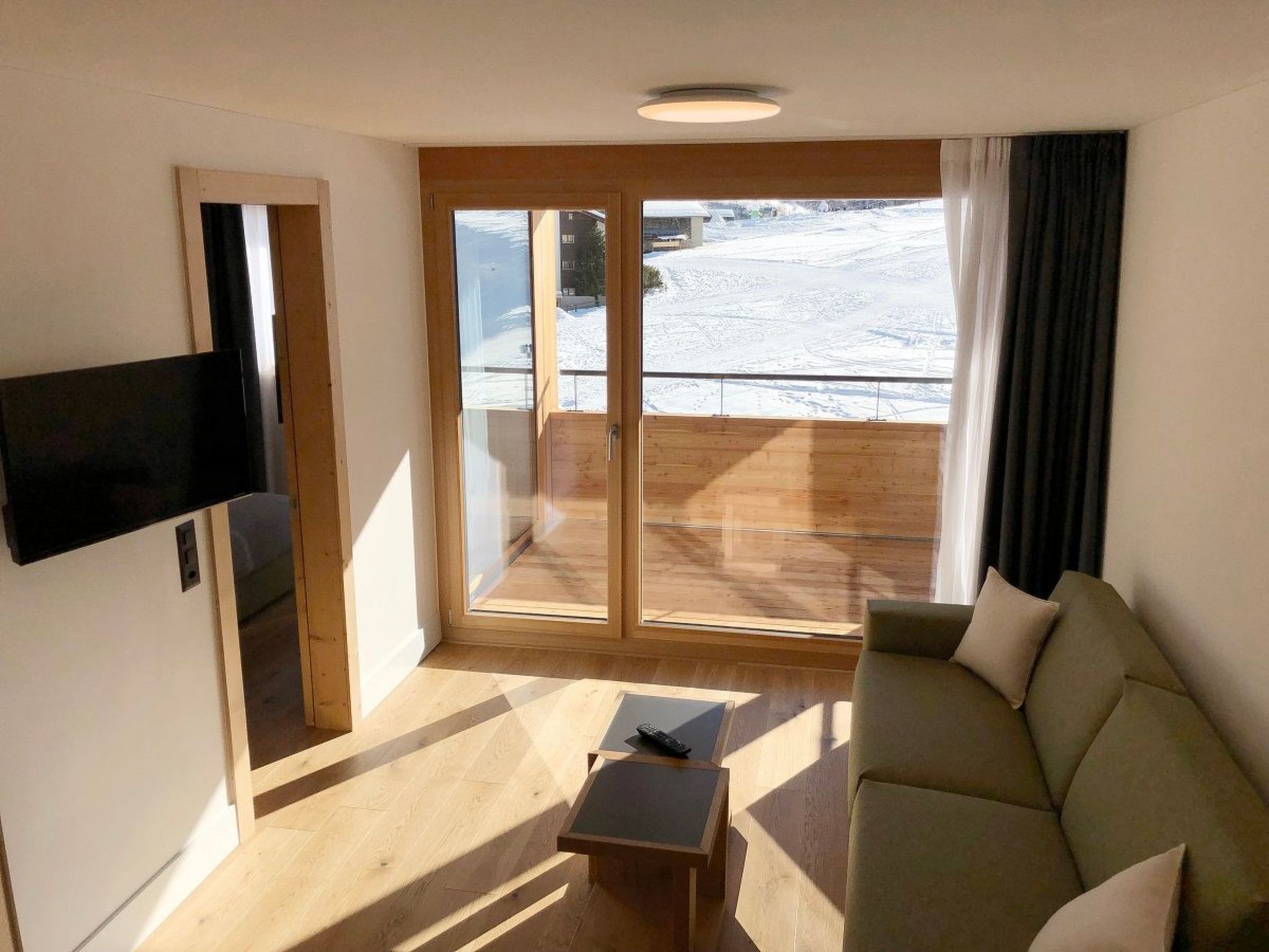 Une pièce comprenant un canapé, une petite table, un téléviseur fixé au mur et une porte-fenêtre coulissante donnant sur un espace extérieur enneigé.