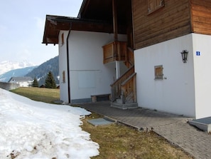 A building with a wooden staircase leading to a landing, a white exterior wall, and a view of snow-covered ground and mountains in the background. - Londadusa OG, (Rueras/Sedrun), 178.01 by Interhome (Rueras Dieni)